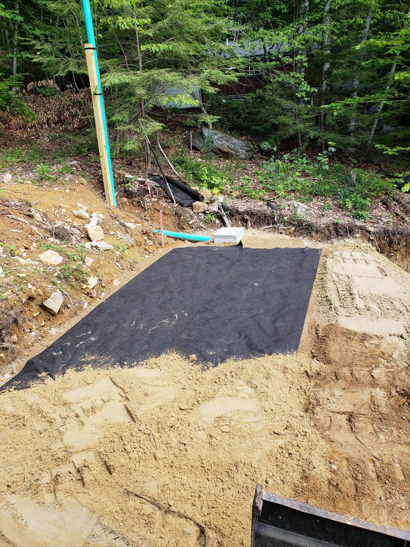 A sandy construction site with black fabric laid on sand. Green pipes and trees in the background.