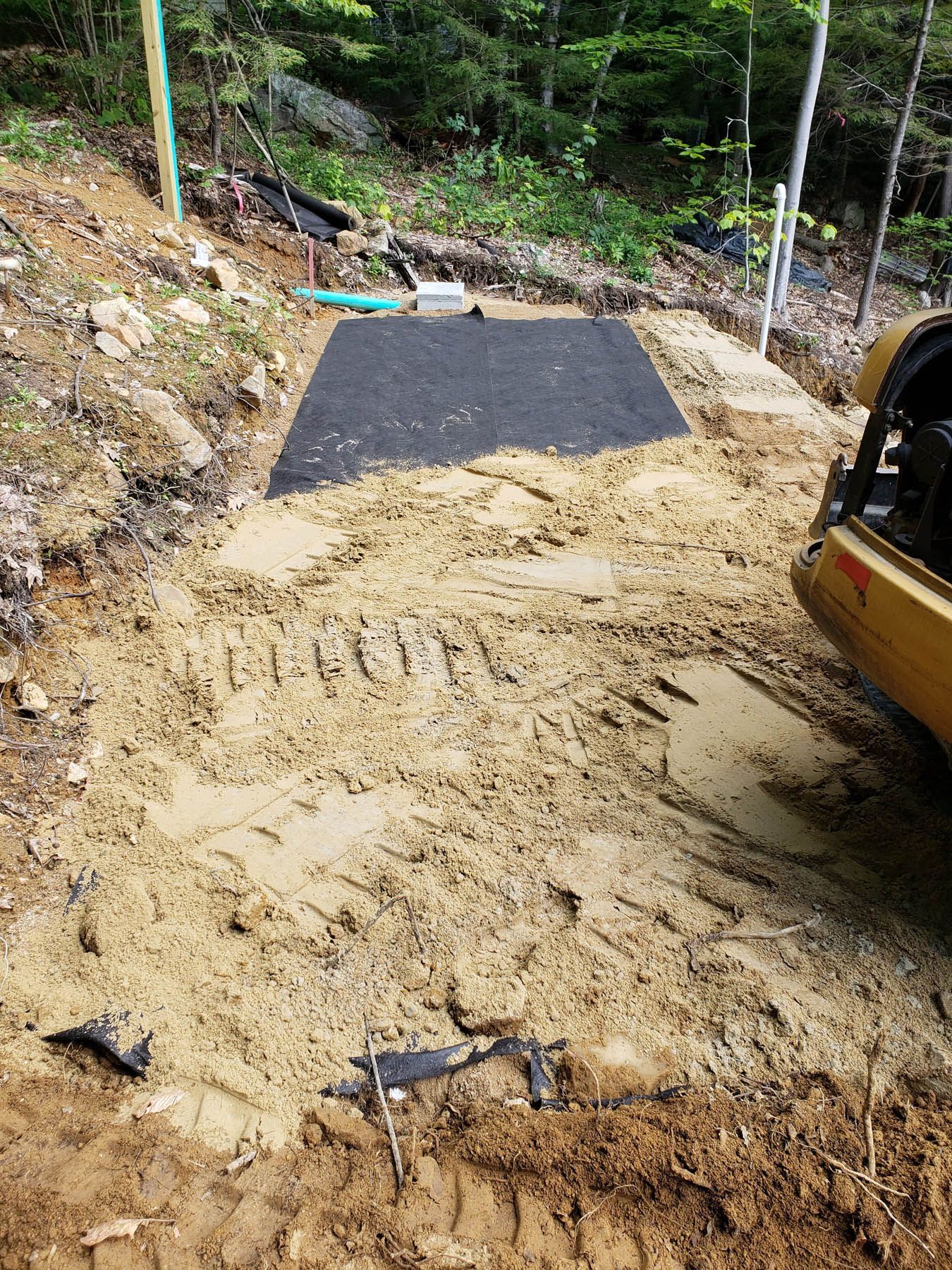A muddy construction site with a black mat and a yellow vehicle, set in a wooded area.