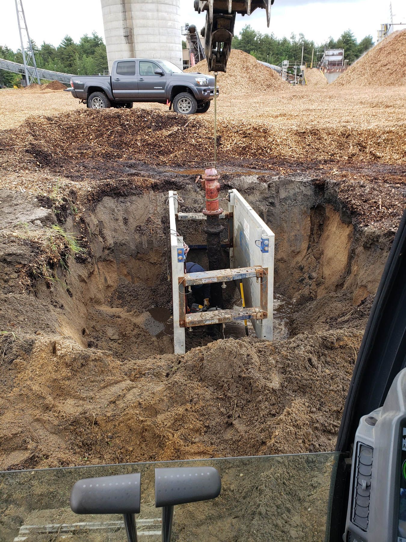 Excavated water valve in a trench, with a truck and wood chips in the background.