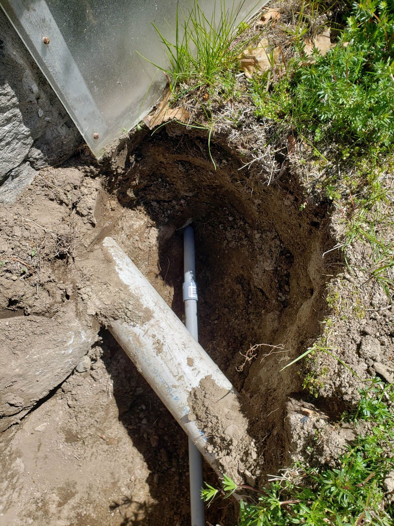 Hole in the ground revealing buried pipes near a structure with a green grassy border.