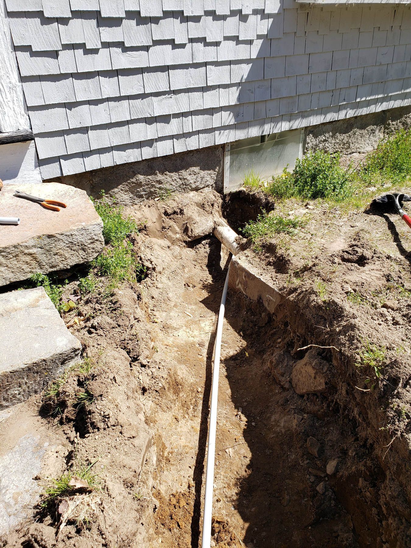 Trench dug next to a building with a white pipe running through it; tools and steps nearby.