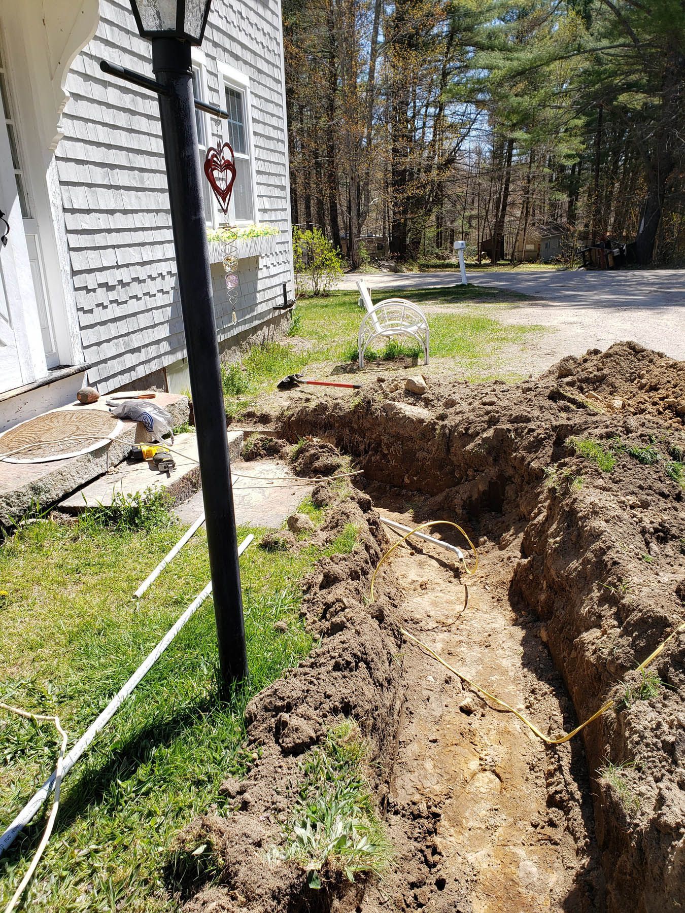 A trench dug near a house for underground utility lines. Dirt, grass, and a black lamp post are visible.
