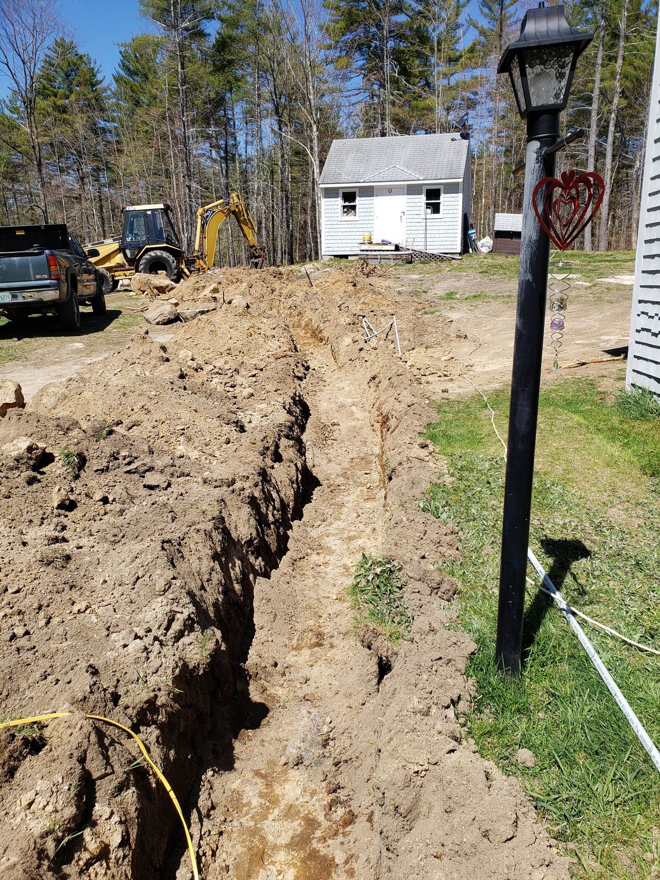 Trench dug in a yard; excavator and truck visible in the background; house in the distance.