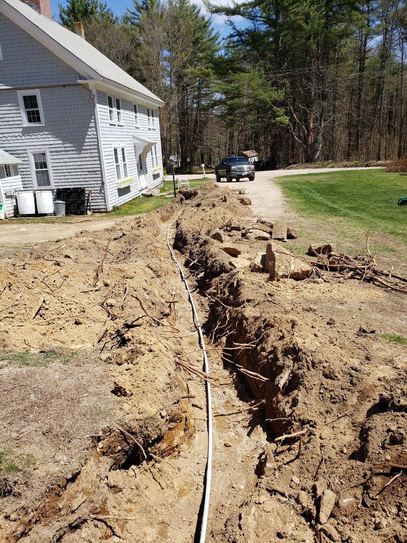 A trench dug in the dirt, with a white pipe, next to a house and road, with a car in the distance.