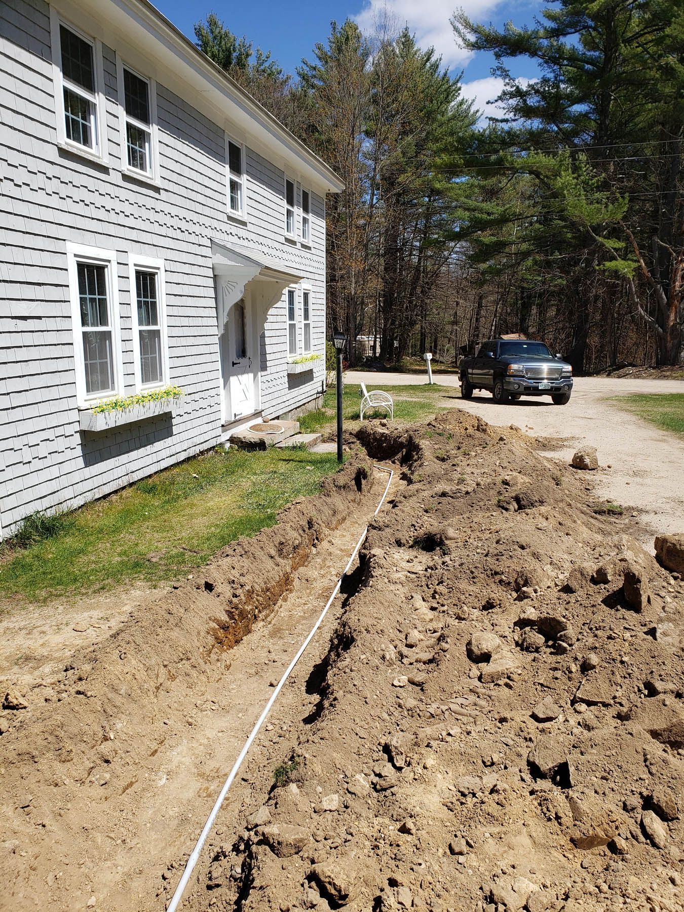 A long trench next to a house, with a white pipe inside. A truck is parked nearby on a dirt road.