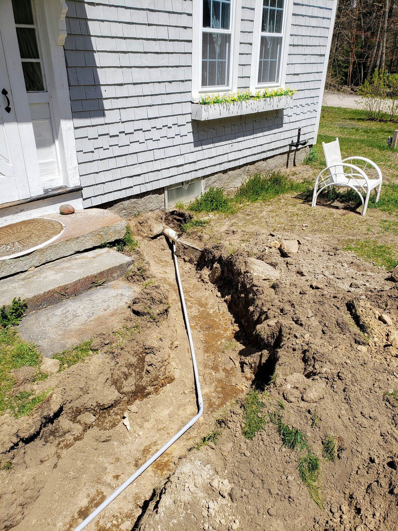 Trench dug along a house foundation, white pipe inside. House and steps in view.