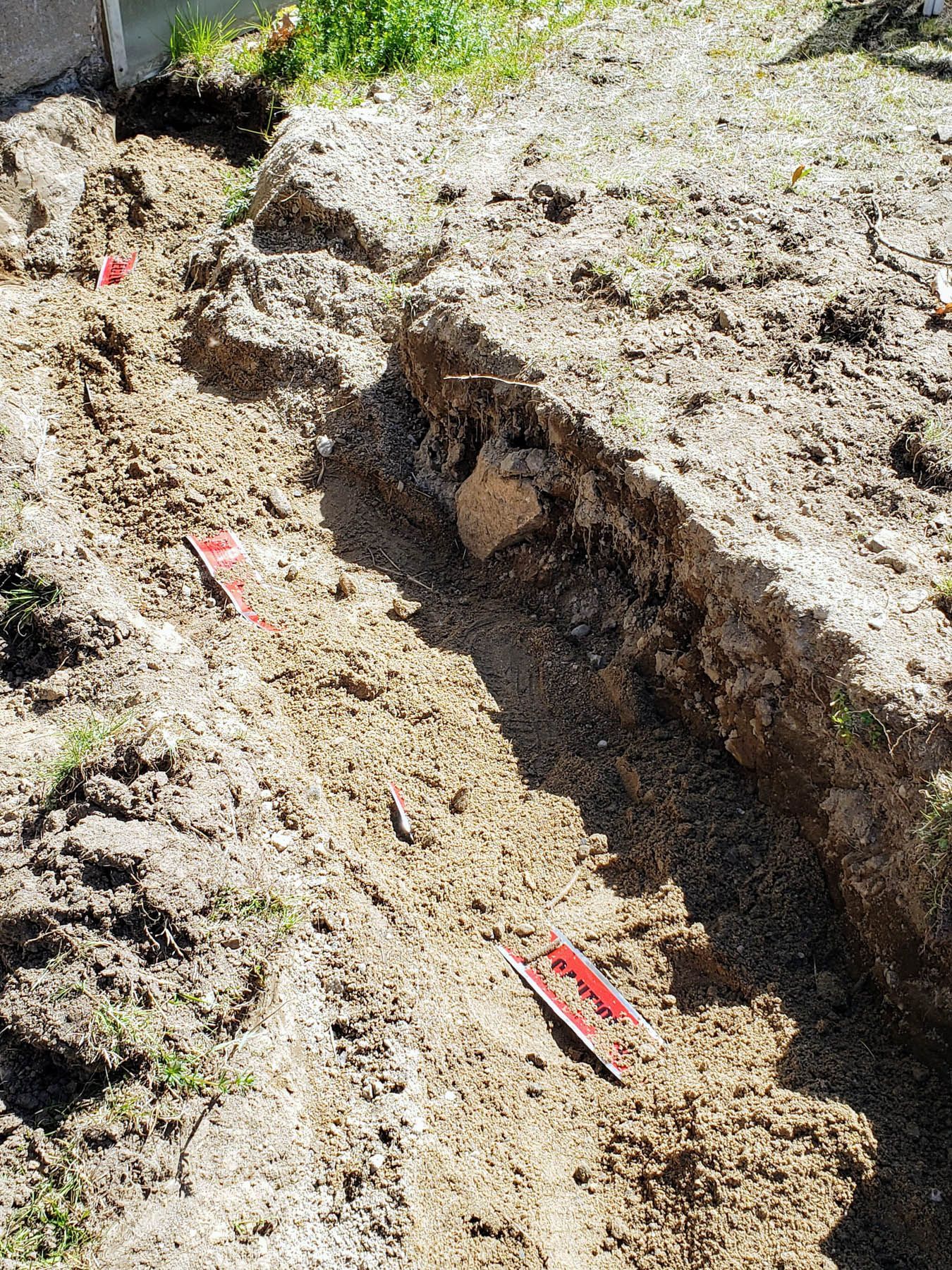 A narrow trench dug in brown soil, marked with red markers, in an outdoor setting.