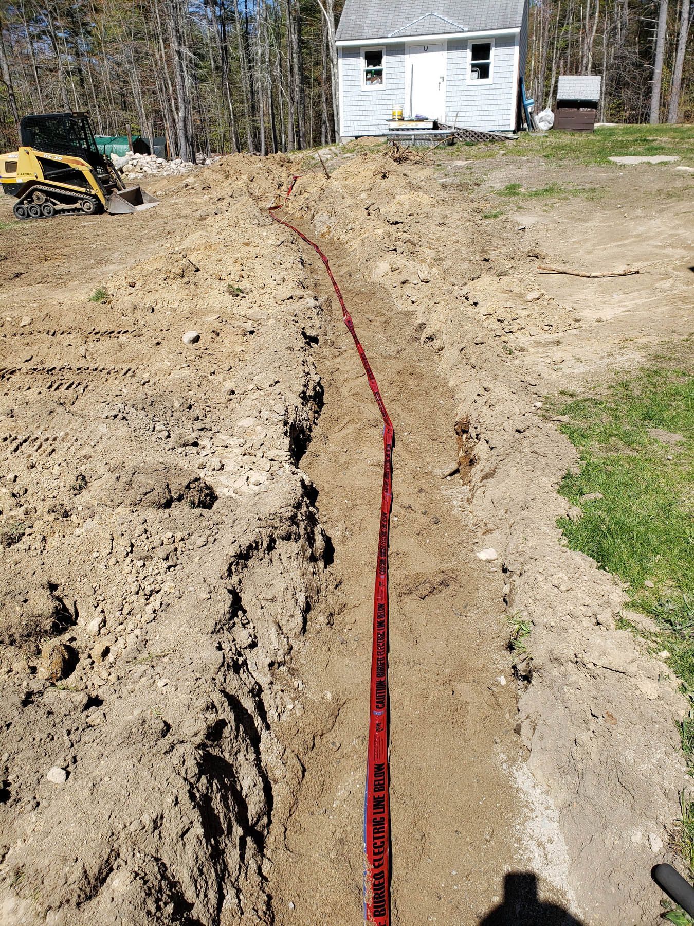 A trench dug in a yard, with a red measuring tape inside. A small building sits in the background.