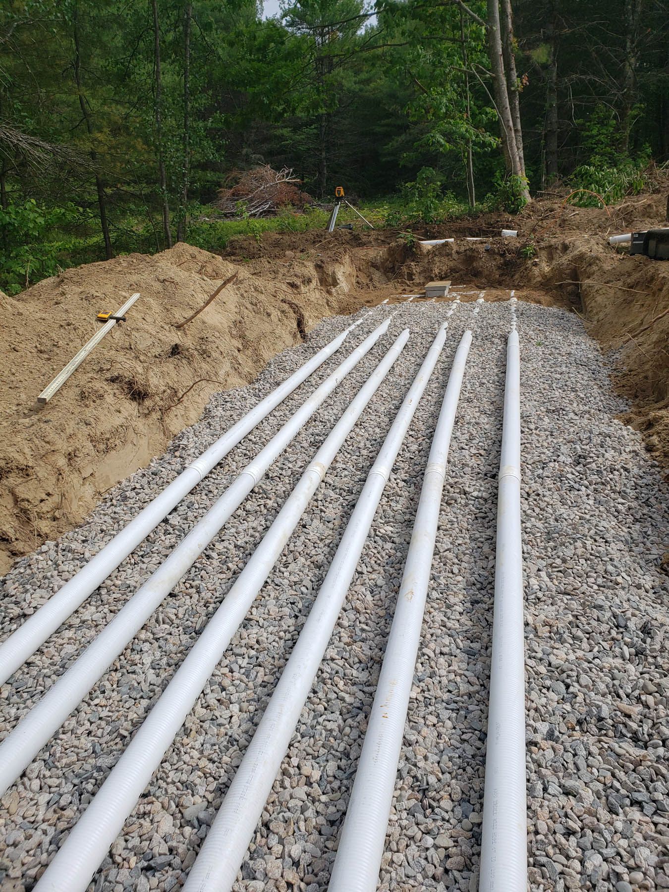 Six white perforated pipes laid in gravel, part of a septic system, outdoors in a trench.