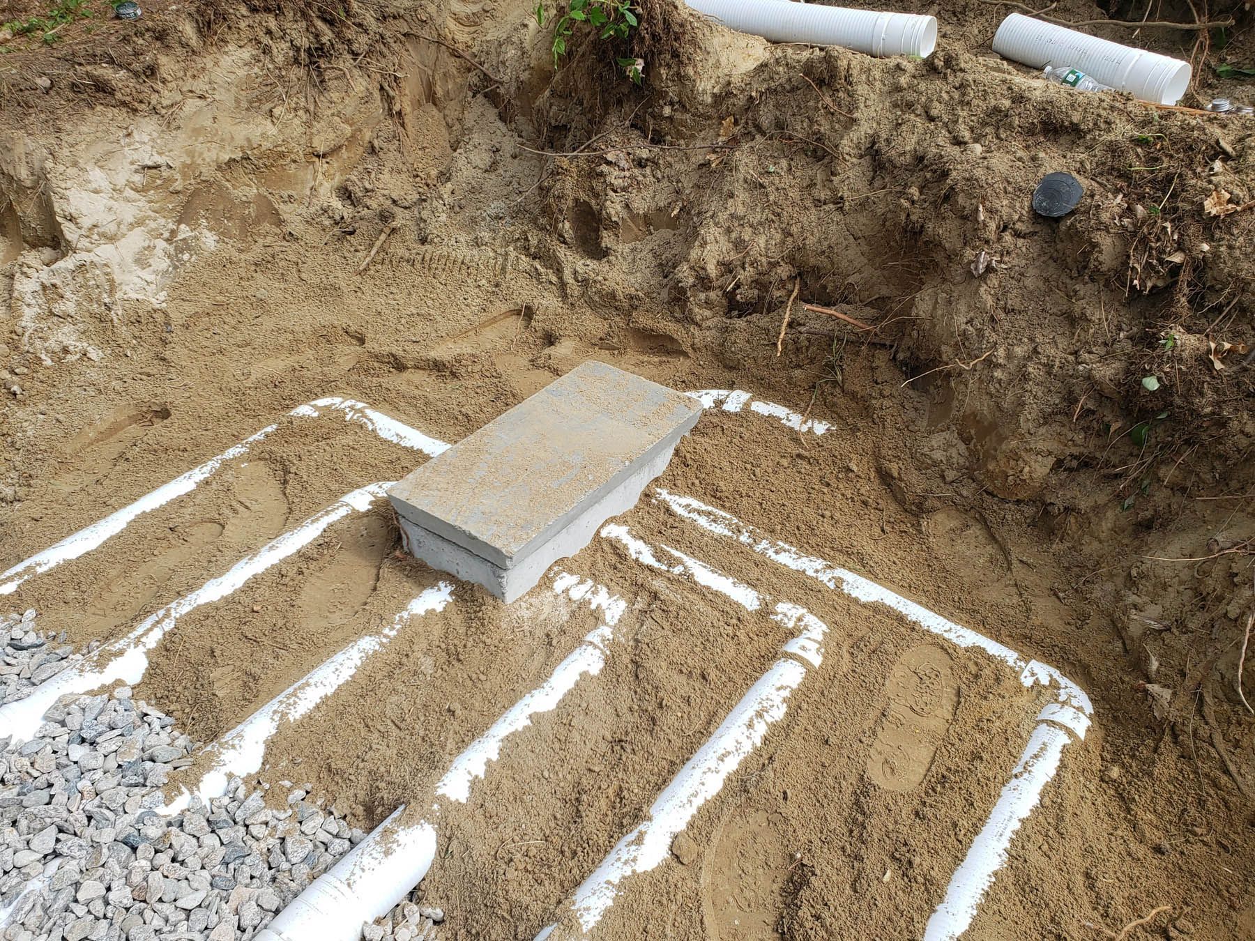 Excavation with white pipes laid in sand for drainage. A brick sits atop the pipes.