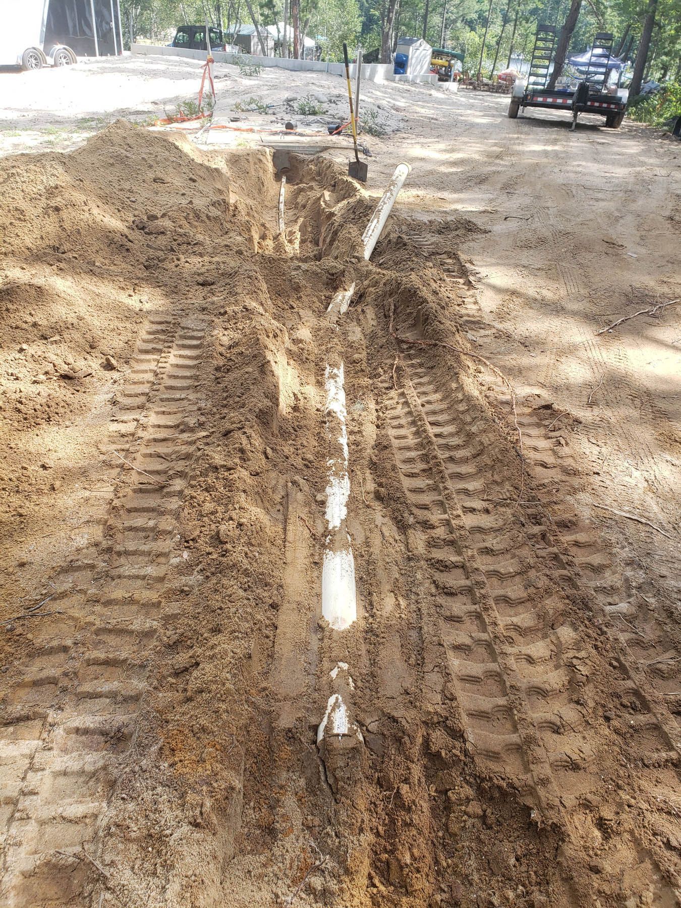 Trench dug in muddy ground with white pipes installed. Construction equipment and a structure are visible in the background.