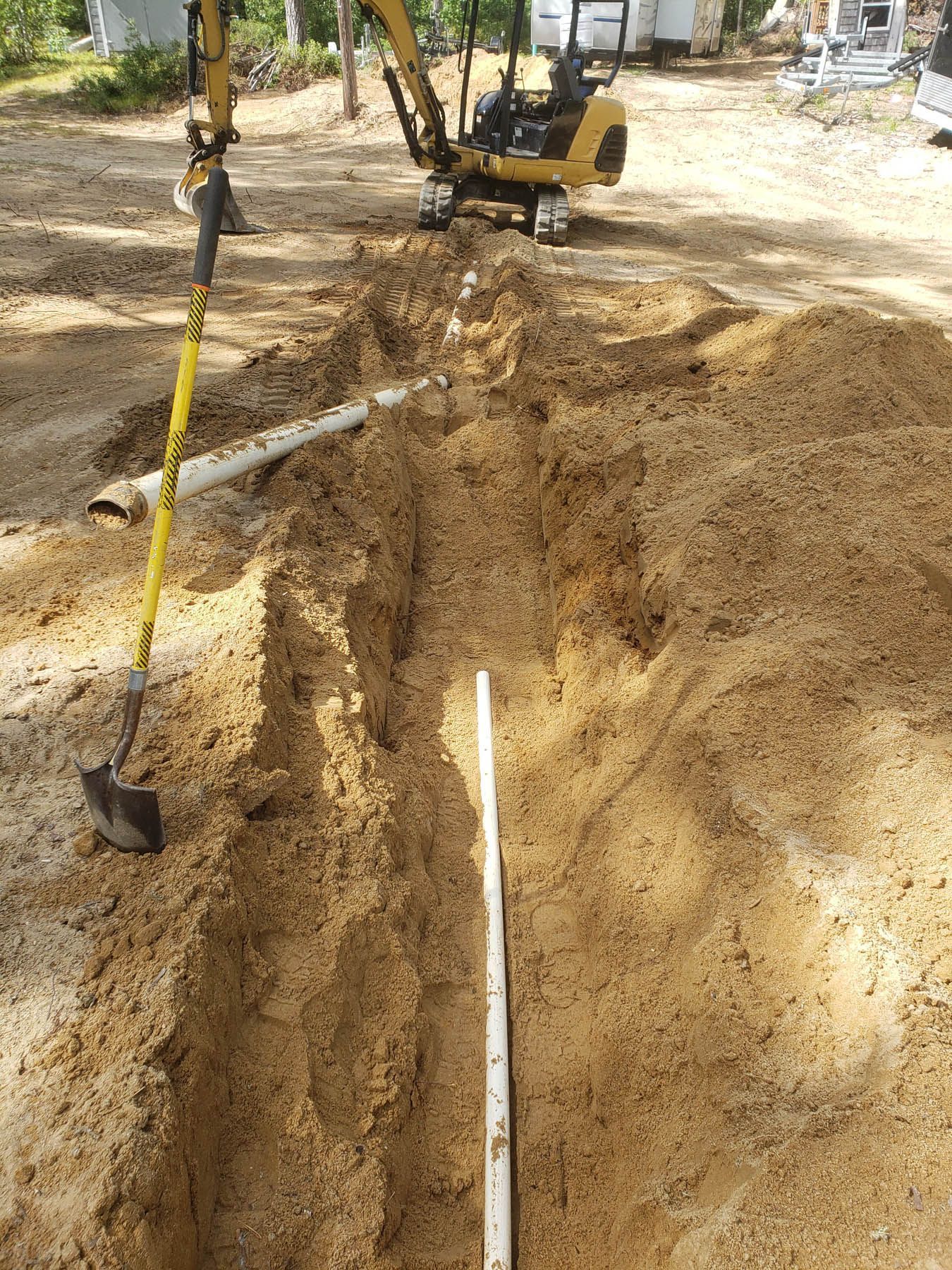 A small excavator digs a trench in sand. A shovel and white pipe are in the trench.