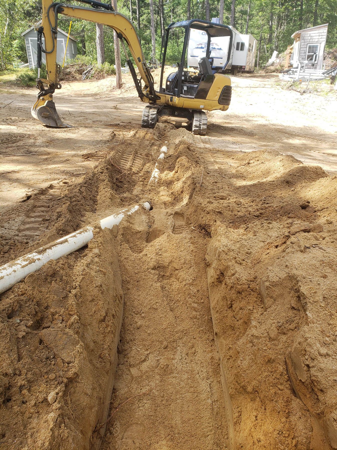 Excavator digging a trench with white pipe laid inside. Construction site, daylight.
