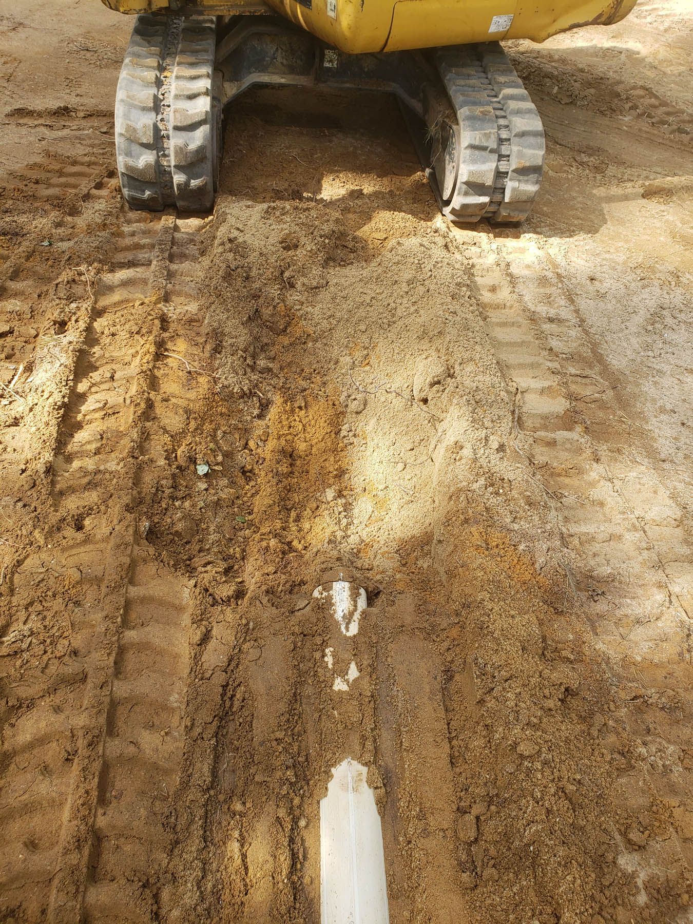 Excavator tracks over freshly dug trench, revealing a white pipe in the sand.