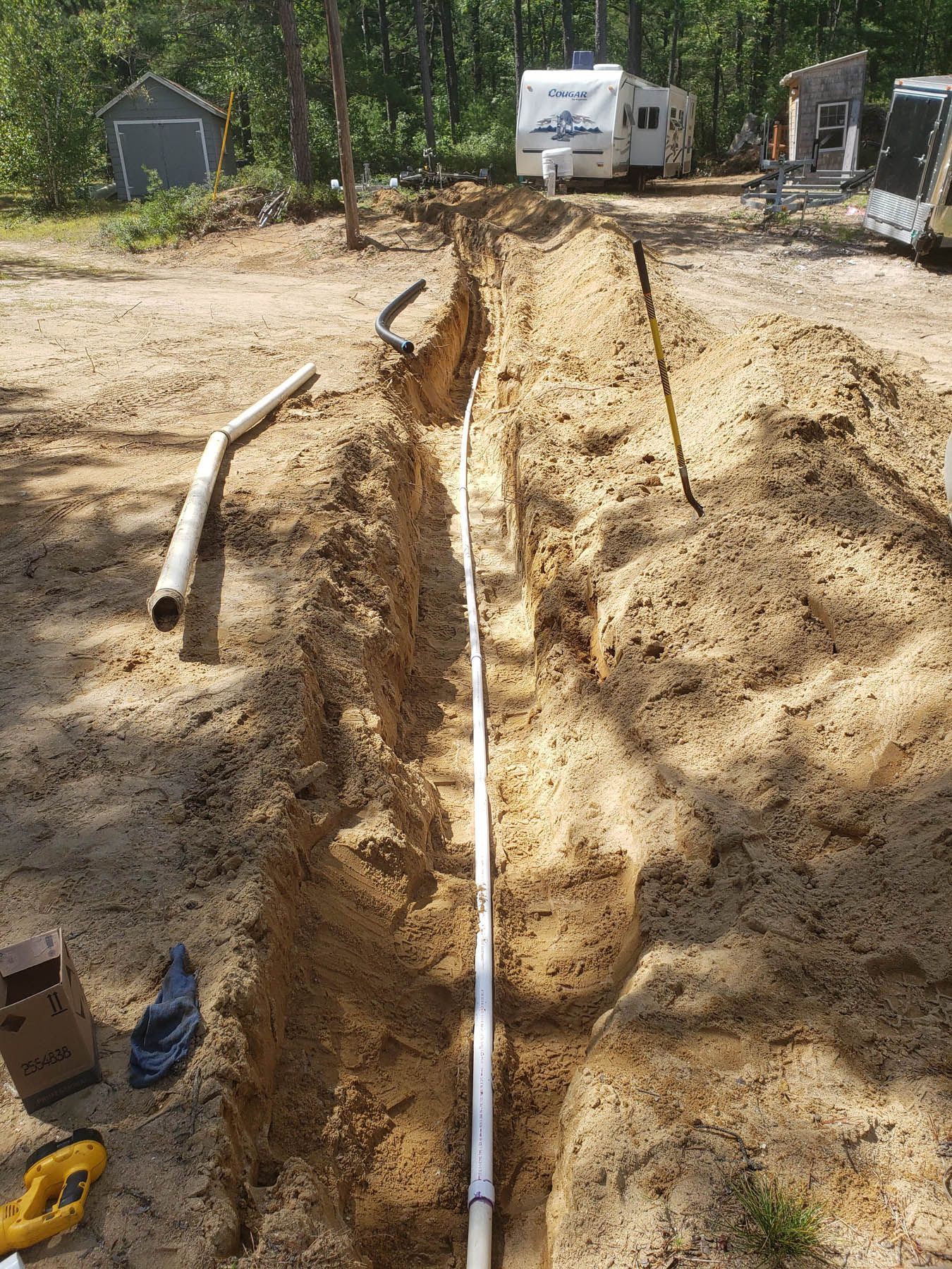 A long trench with white pipe laid in it, outdoors with a trailer and trees in the background.