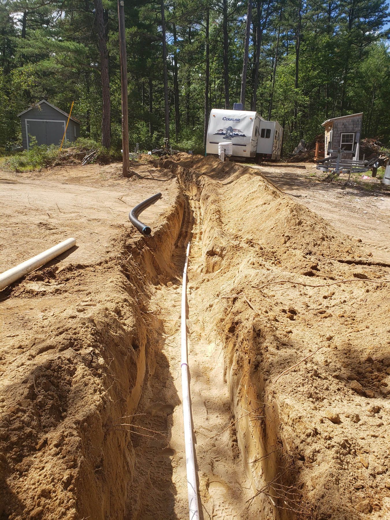 Trench dug in dirt with white and black pipes, a camper, and trees in the background.