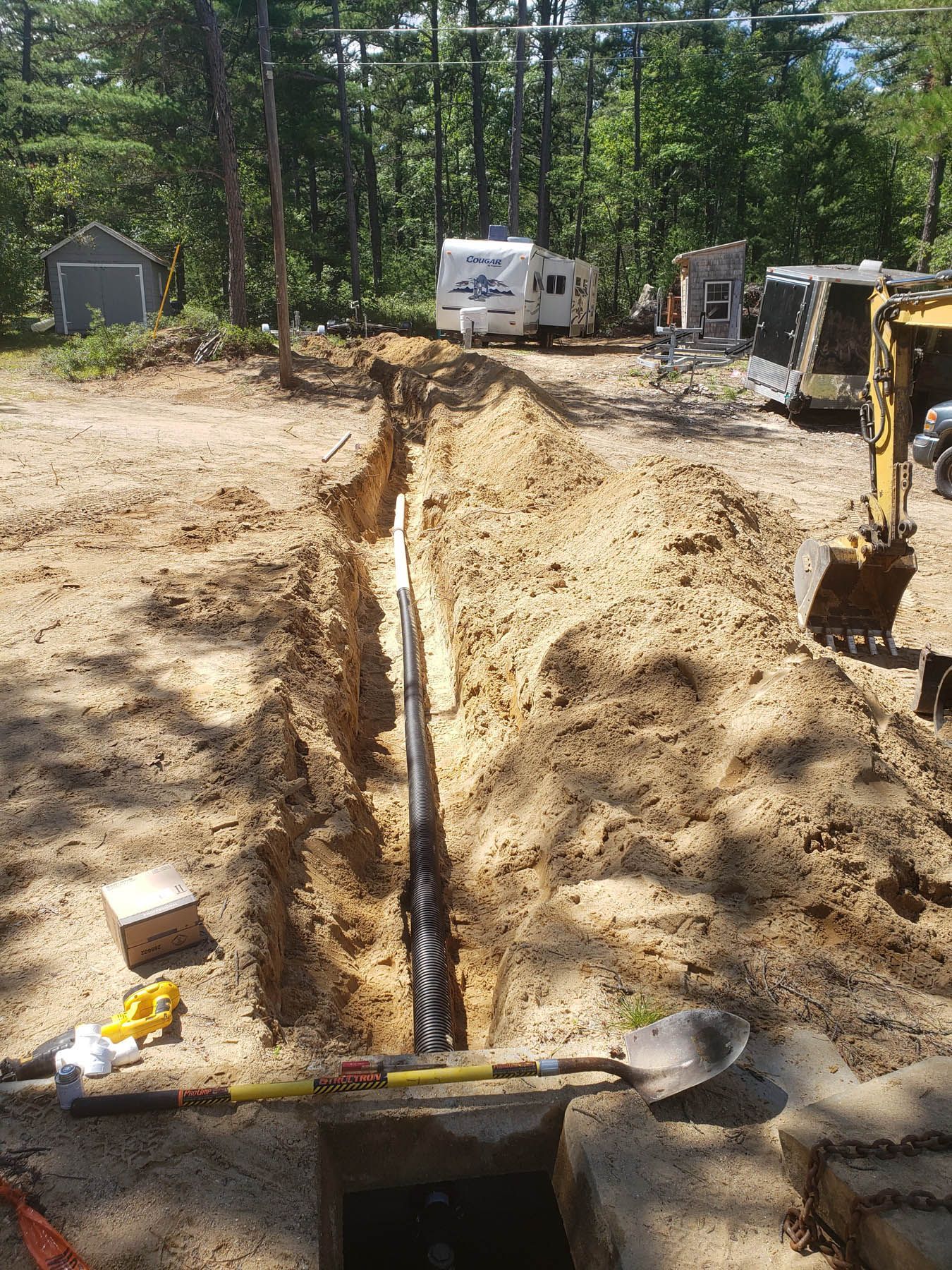 Trench dug in sandy ground for a black pipe installation; a small excavator and RV are in the background.