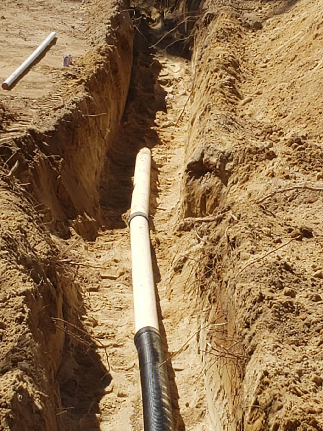 A long, narrow trench with a white pipe in the center; dirt walls and another pipe above.