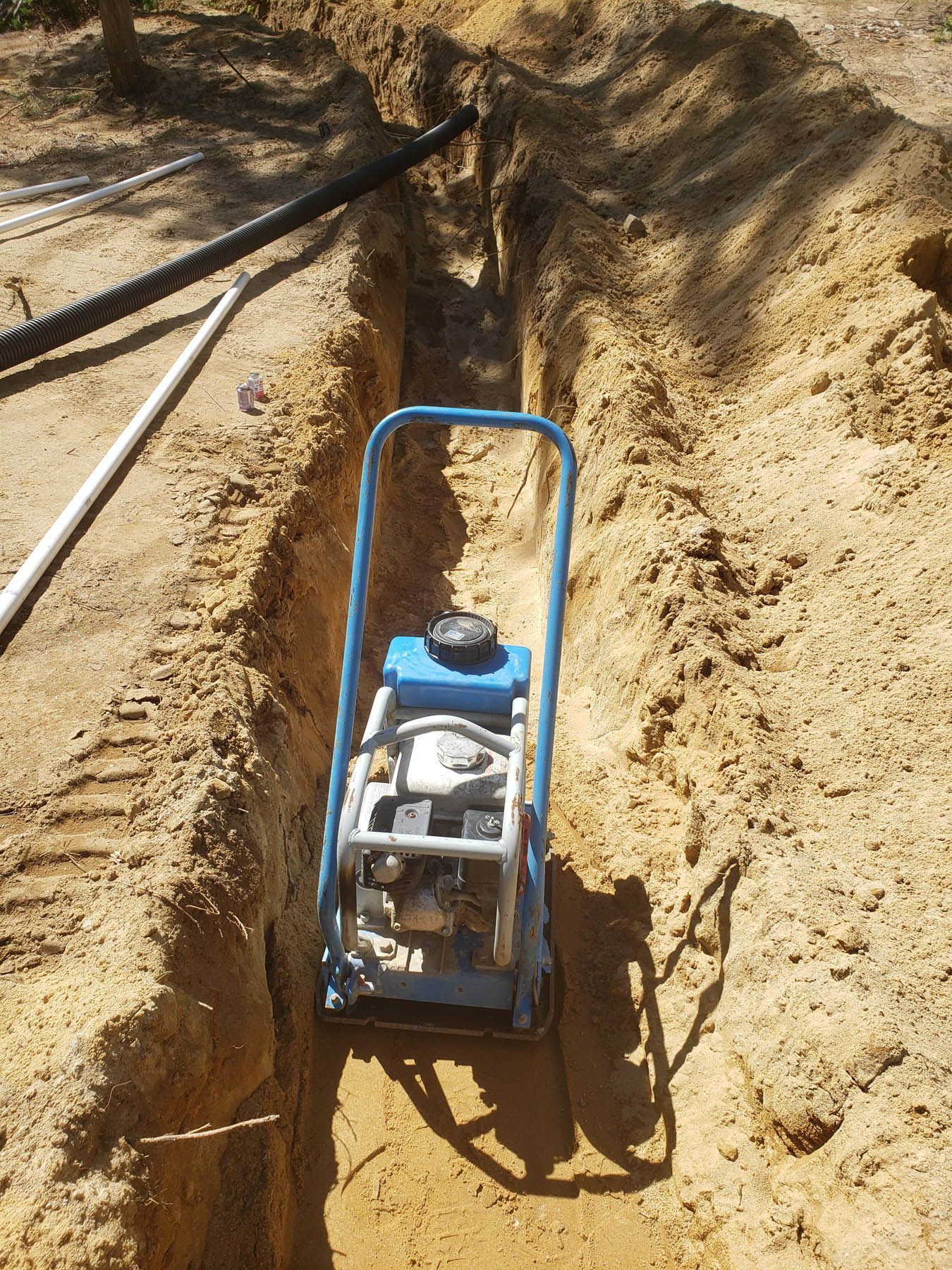 Compaction plate in a sandy trench, with black and white pipes visible, outdoors.