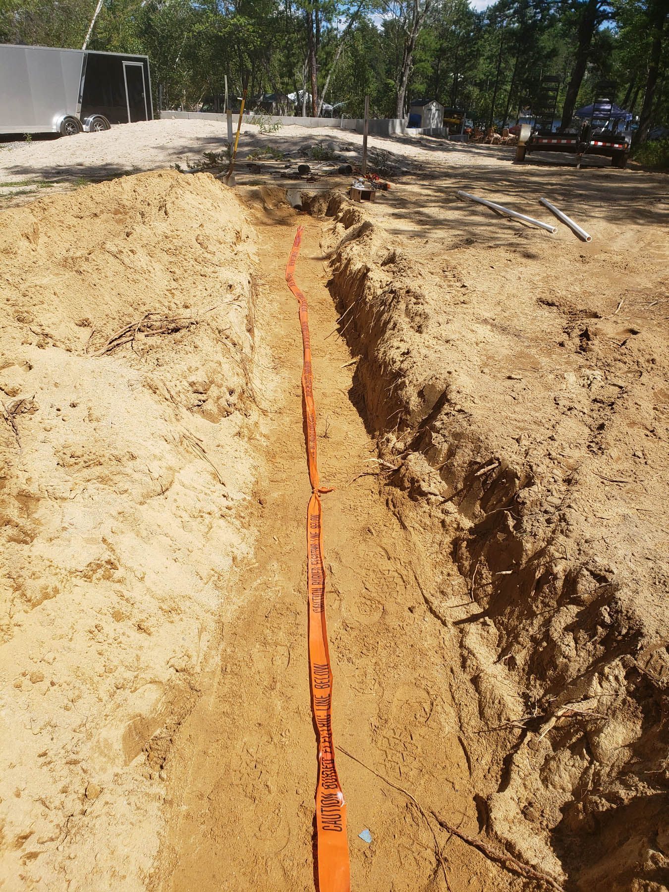 A trench in sandy soil with an orange cable running through it. Construction site in the background.
