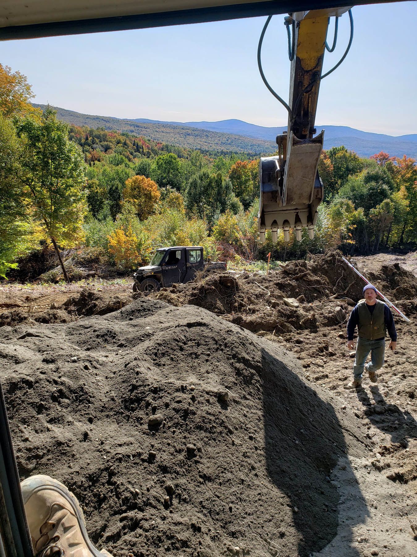 Construction site: excavator, utility vehicle, person walking. Trees and mountains in the background under a blue sky.