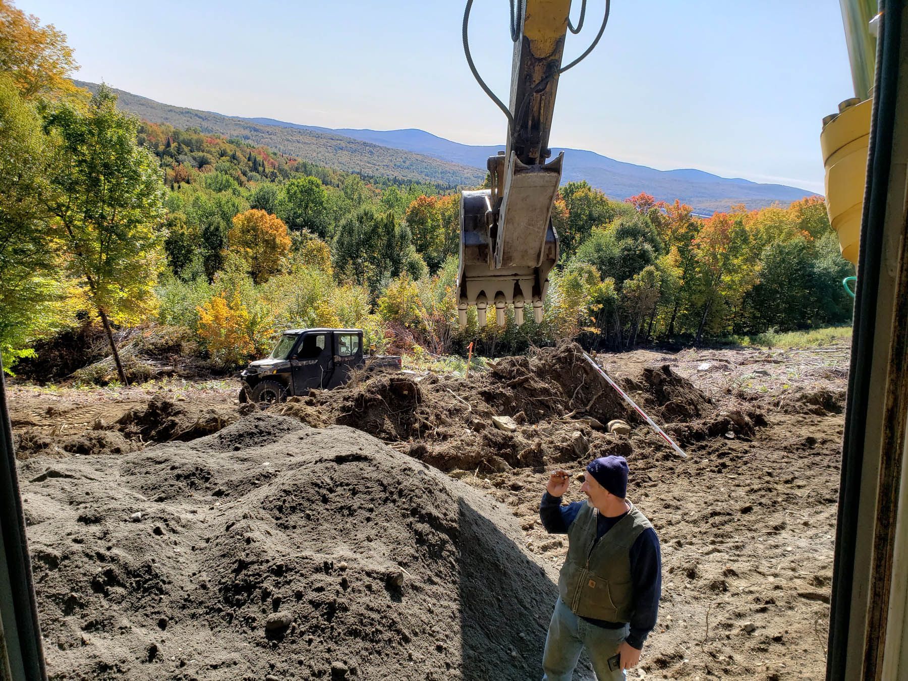 Man observes excavator clearing land near a mountain. A truck and fall foliage visible.