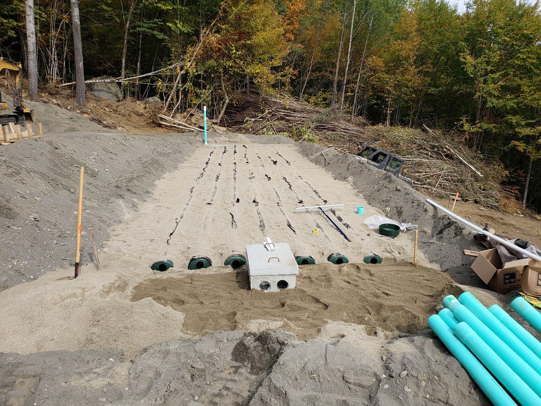 Septic system construction: gravel bed with pipes, distribution box, and forest background.