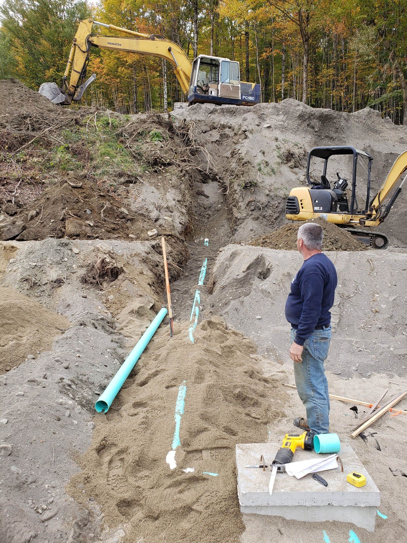 Construction site: man overseeing pipe installation in trench, excavator in background.