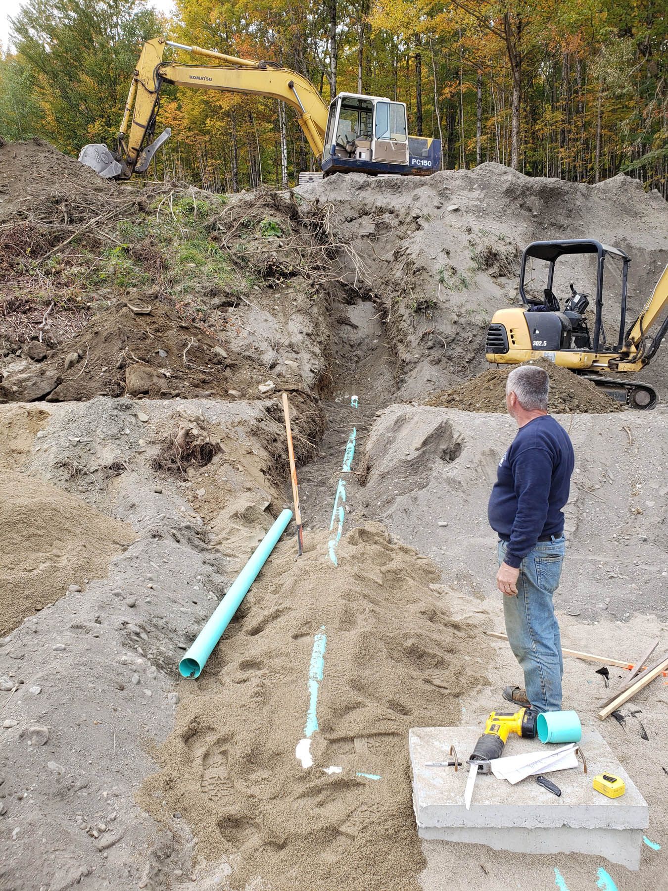 Man observing pipe installation in a trench at a construction site with excavators and dirt.