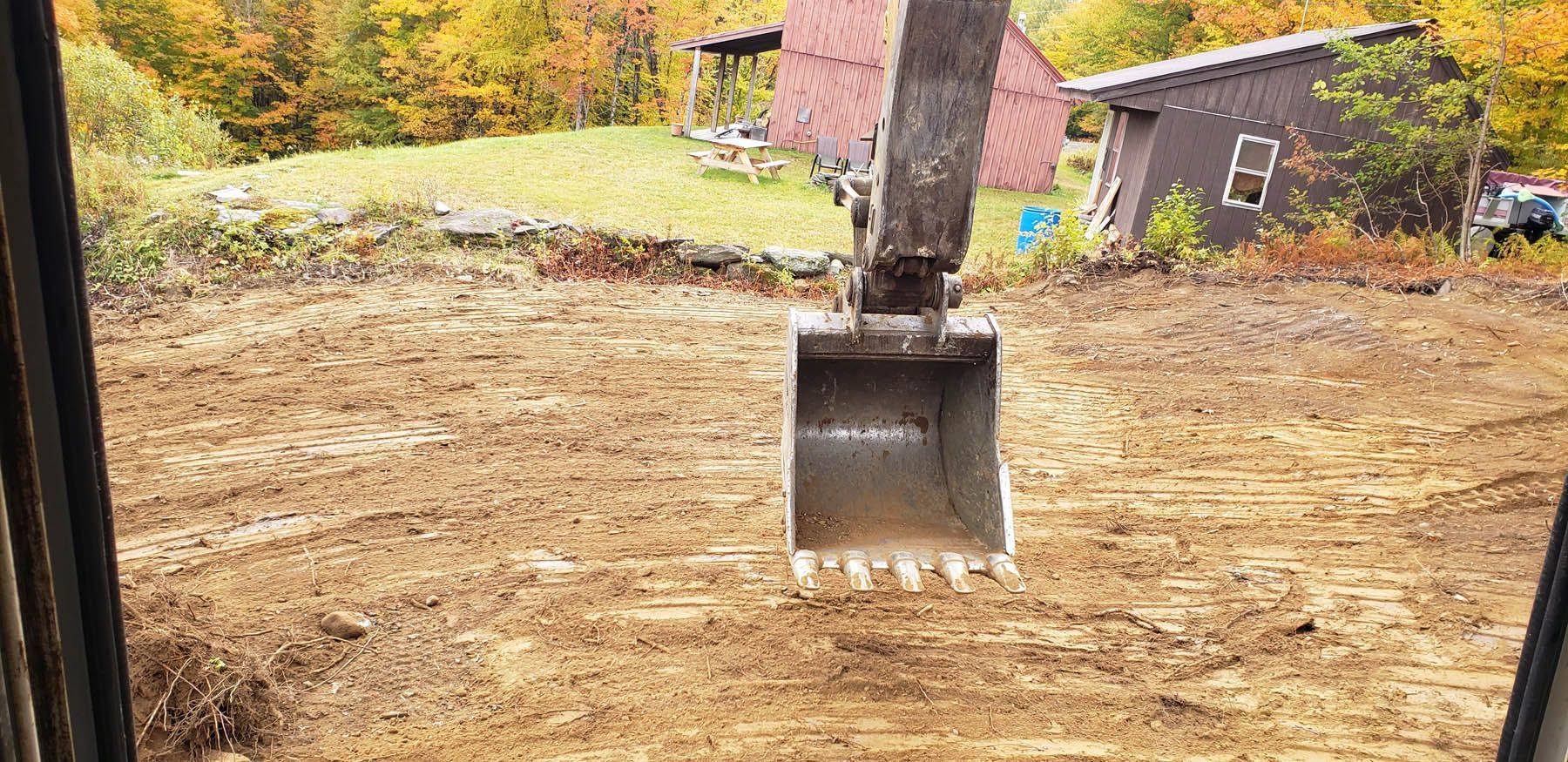 An excavator bucket scoops up dirt in a field, with a house and trees in the background.