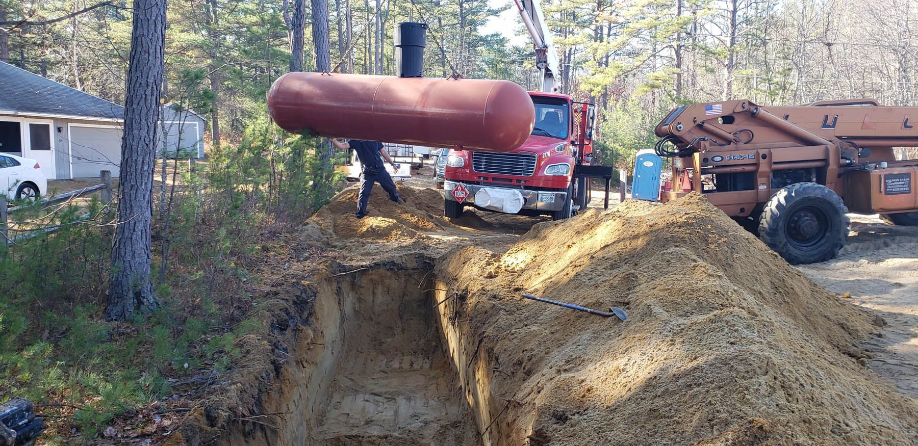 A large propane tank is being lowered into a trench near a house with a red truck and an excavator.