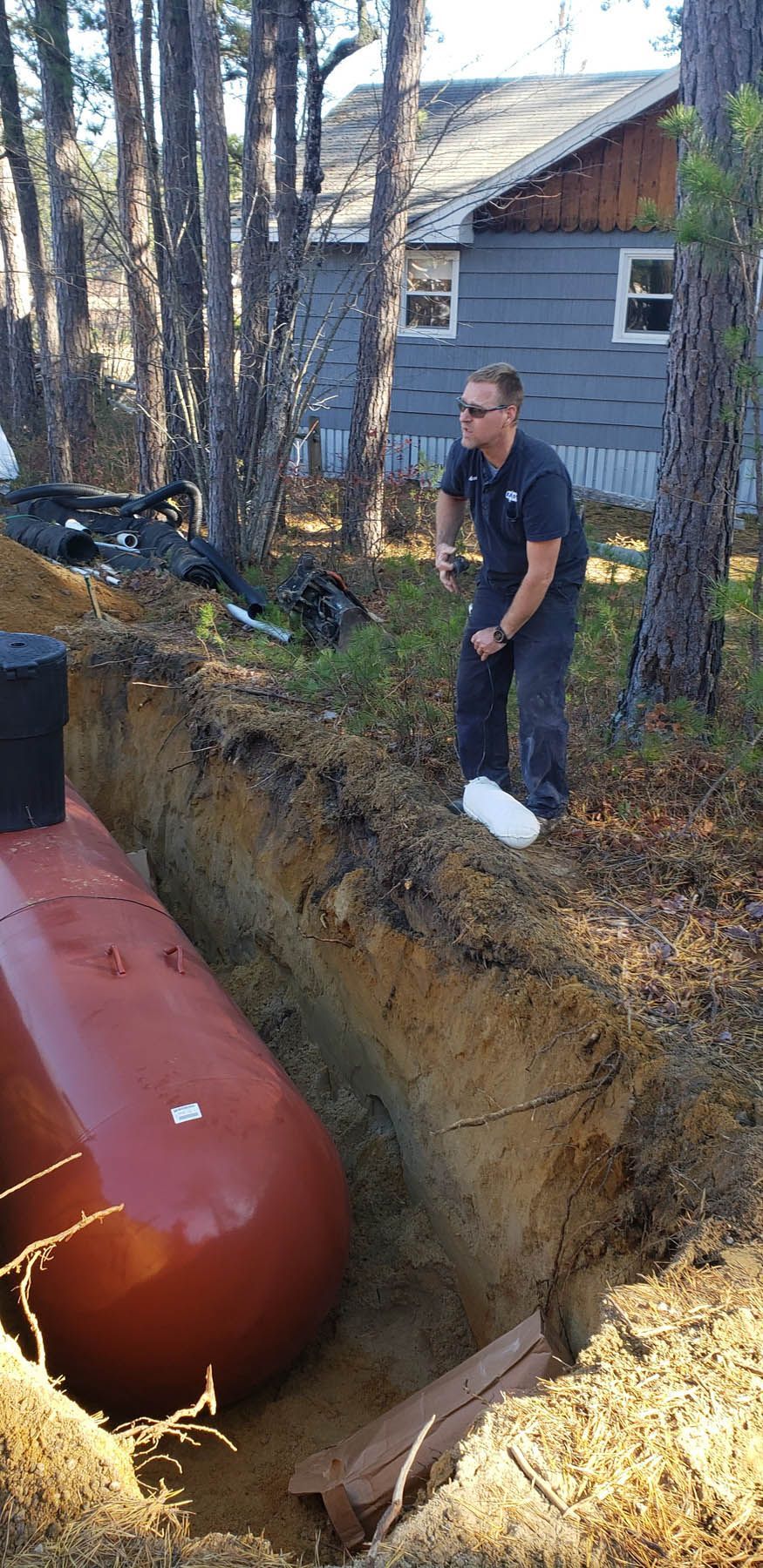 Man in trench inspecting a buried propane tank, with a house in the background.