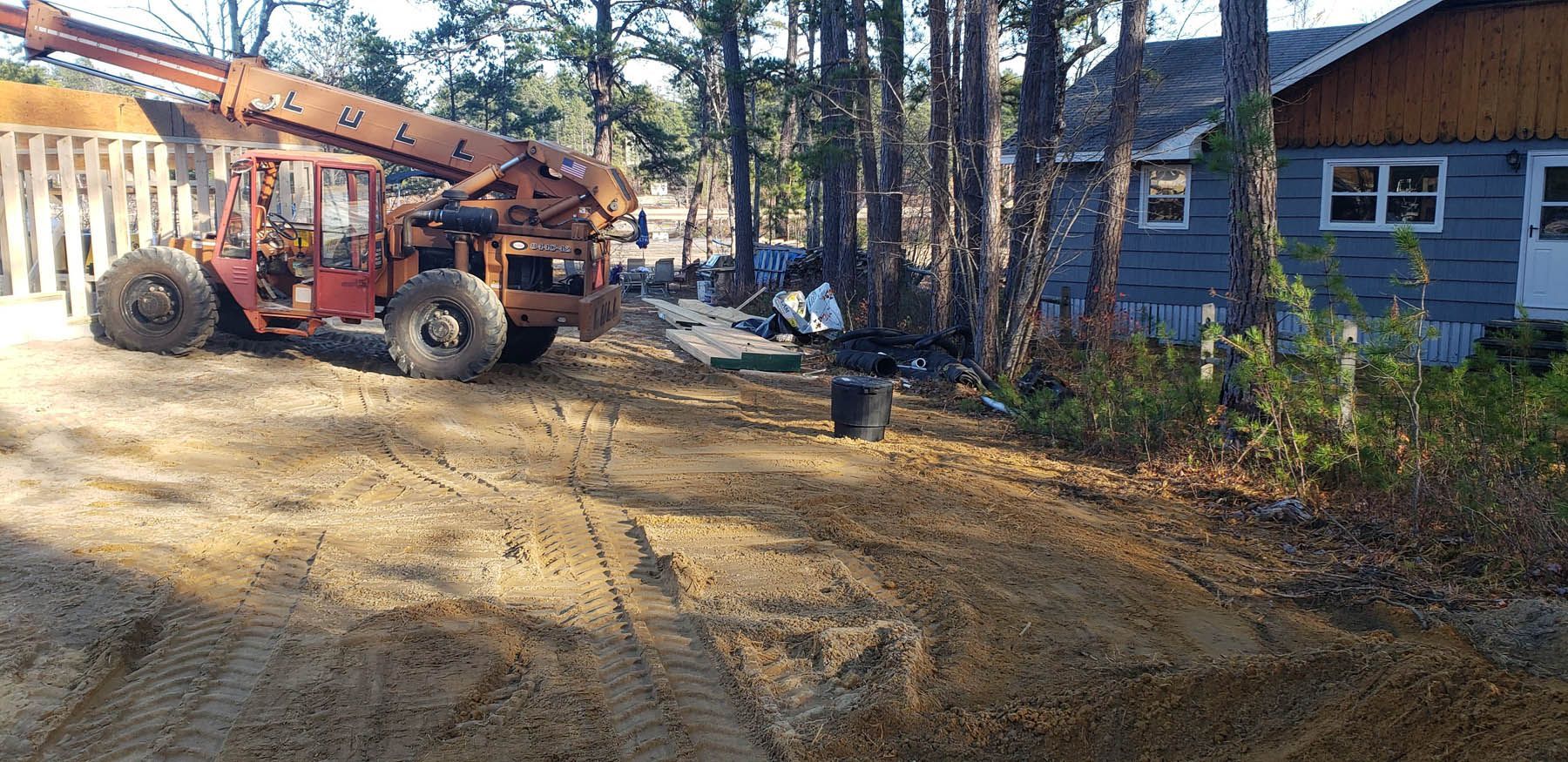 An orange telehandler on a construction site near a blue house, tall trees, and a wooden fence.