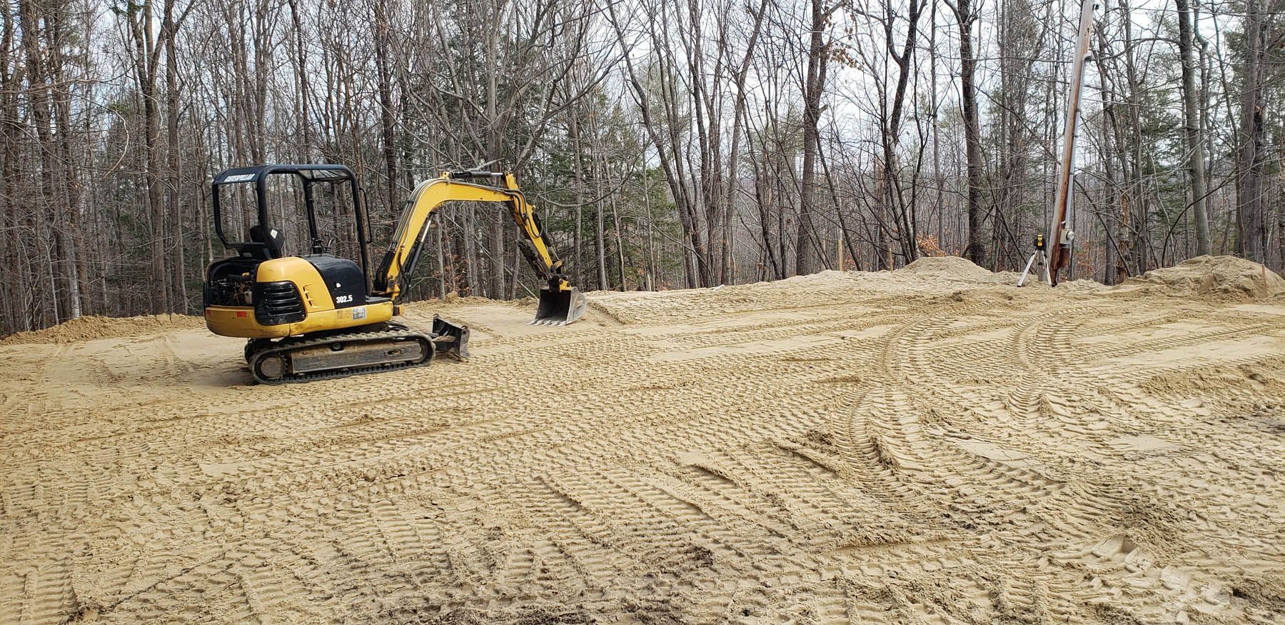 Yellow excavator on a sandy terrain, clearing the ground for construction, with forest background.