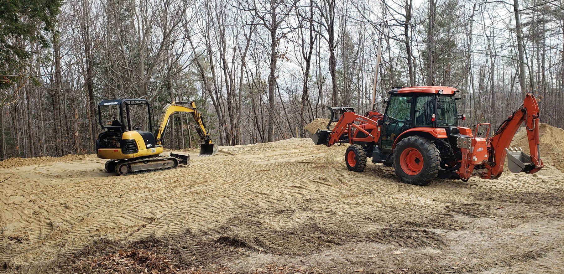 A yellow excavator and orange tractor working in a wooded area, surrounded by mulch.