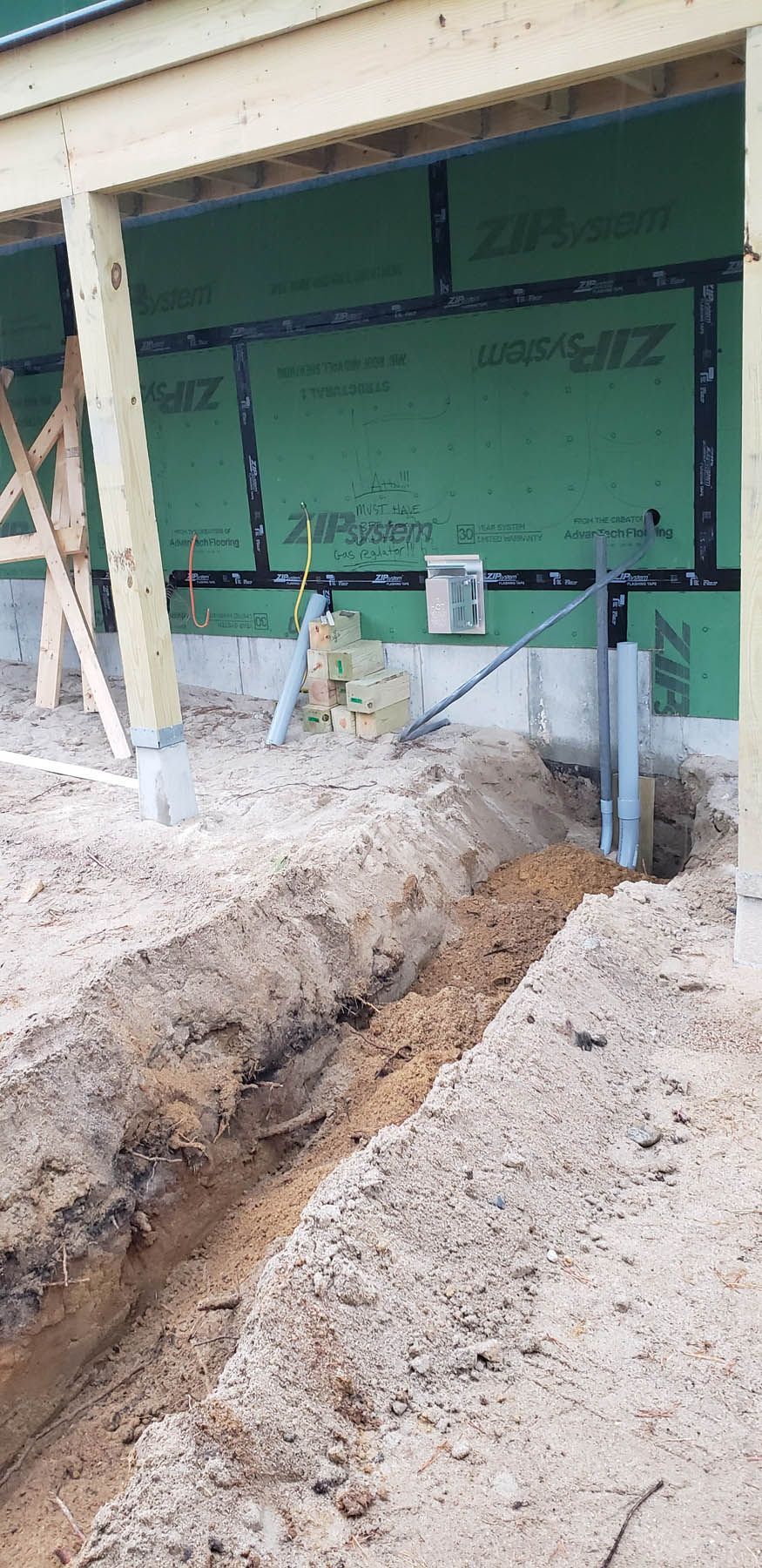 A trench dug in sand with pipes and a building under construction.