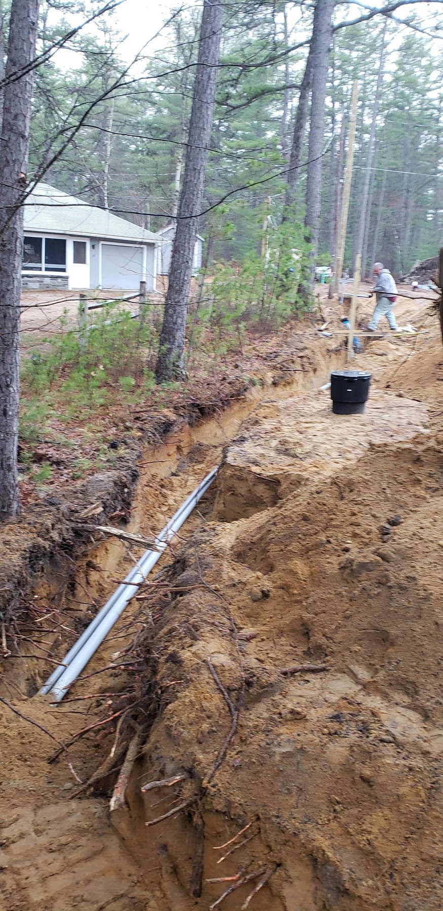 A trench being dug with wires in it near a house, trees, and a person working.