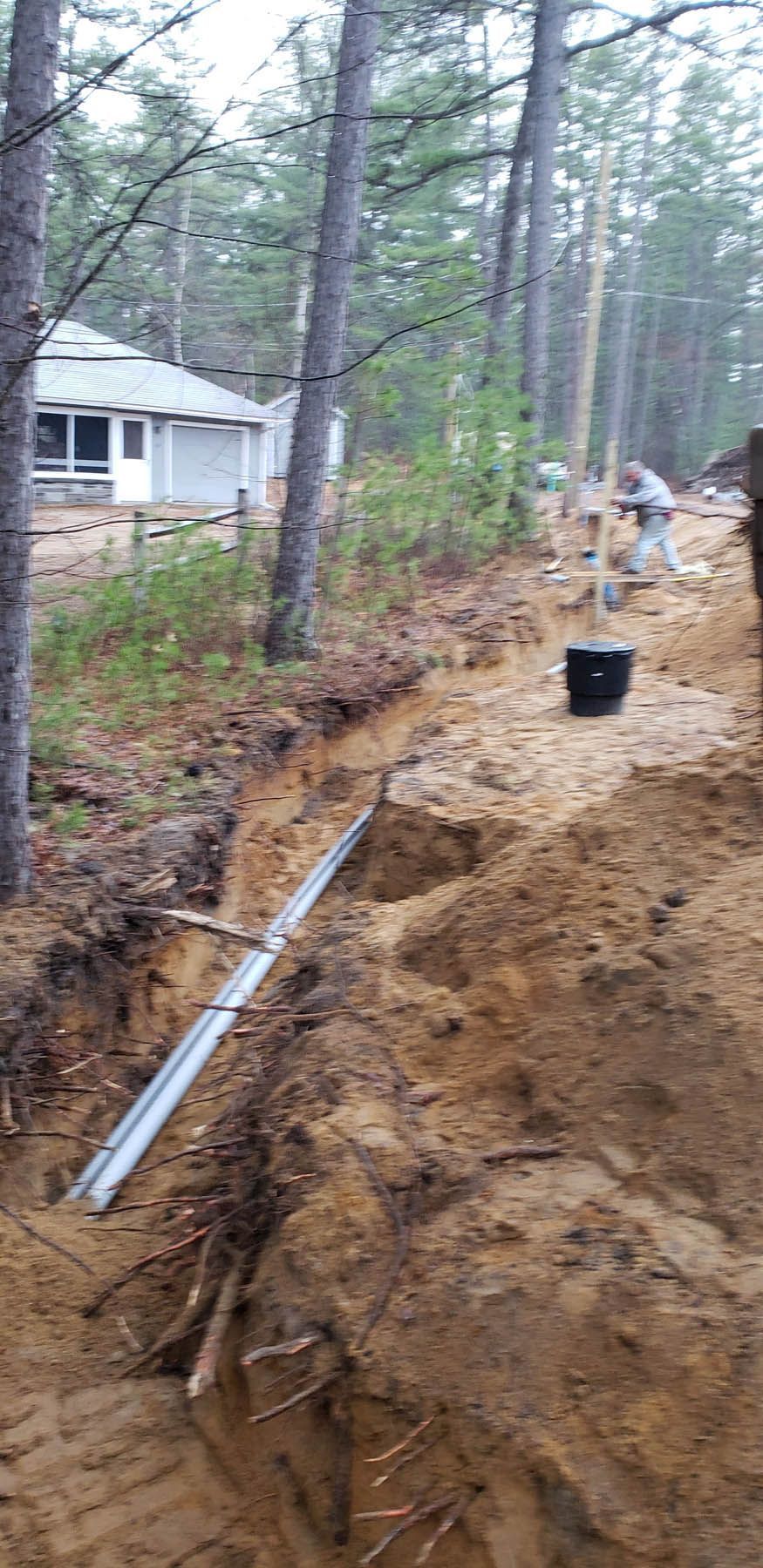 A trench dug along a wooded area for electrical conduit installation. A house is in the distance.