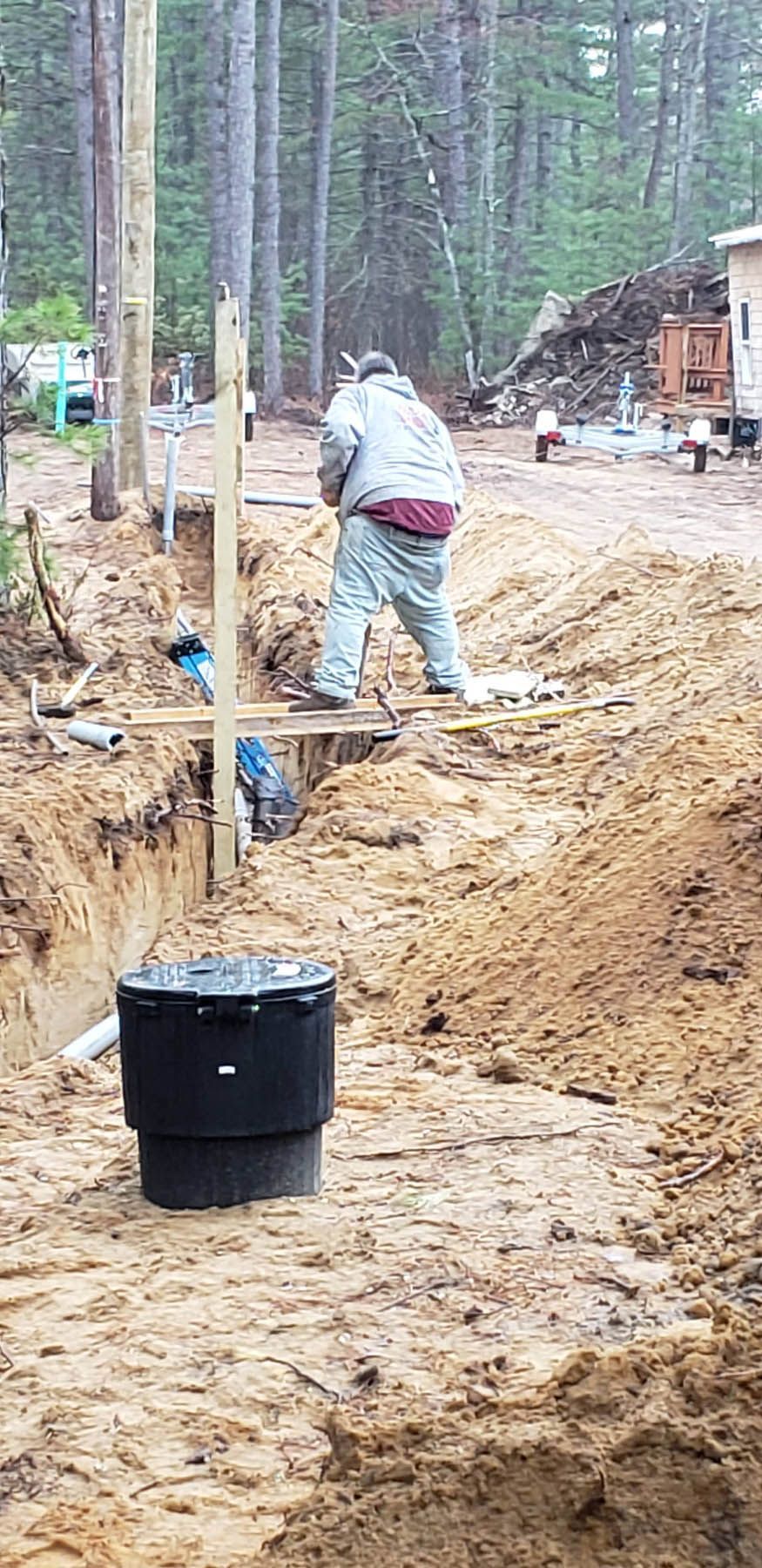 Man working in trench, installing something. Black cylinder in foreground, woods in the background.