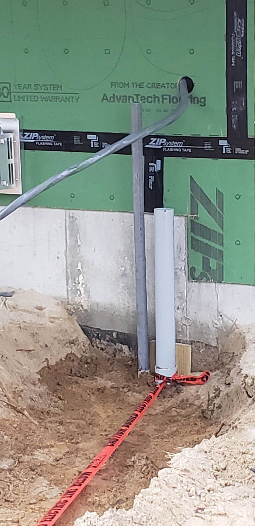 Construction site, gray conduit, and white pipe protruding from the ground near foundation, green wall with black lines and sand.