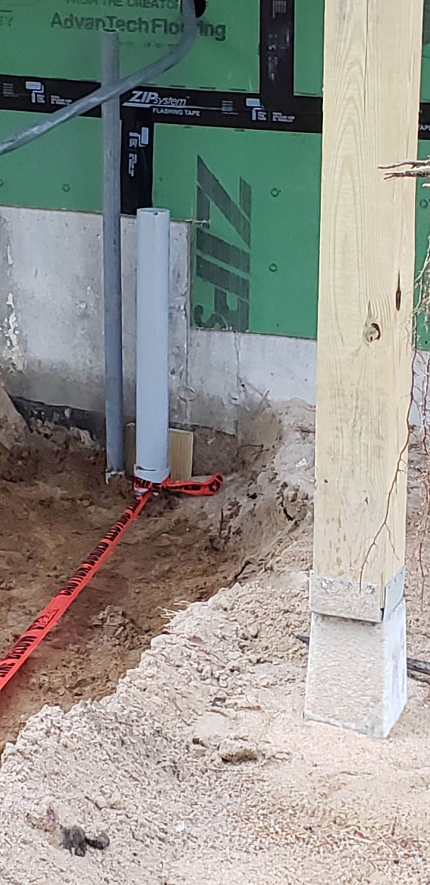 Construction site: exposed foundation with a wooden post, plumbing, and safety tape in the foreground.