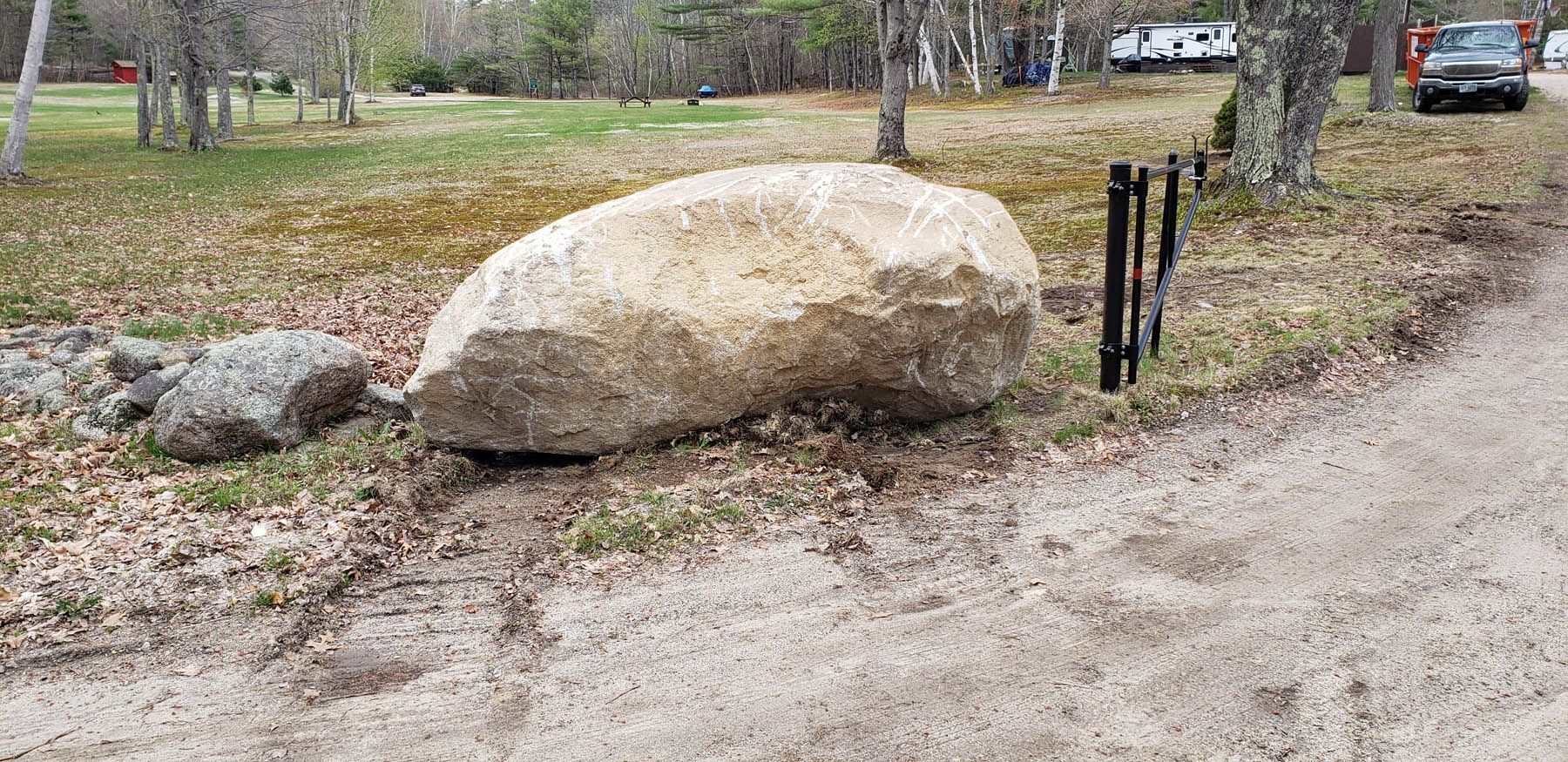 A large, light-colored rock sits next to a dirt road and a few black posts. Green grass and trees are in the background.
