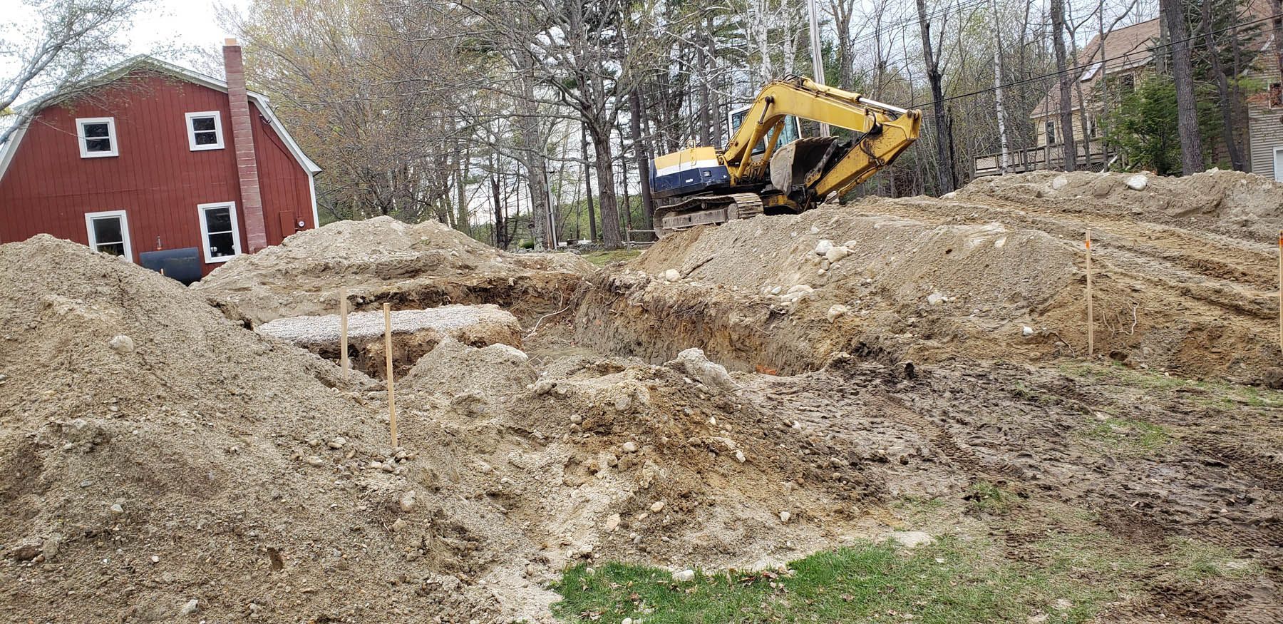 An excavator digging a foundation next to a red house. Earth mounds surround the excavation site.