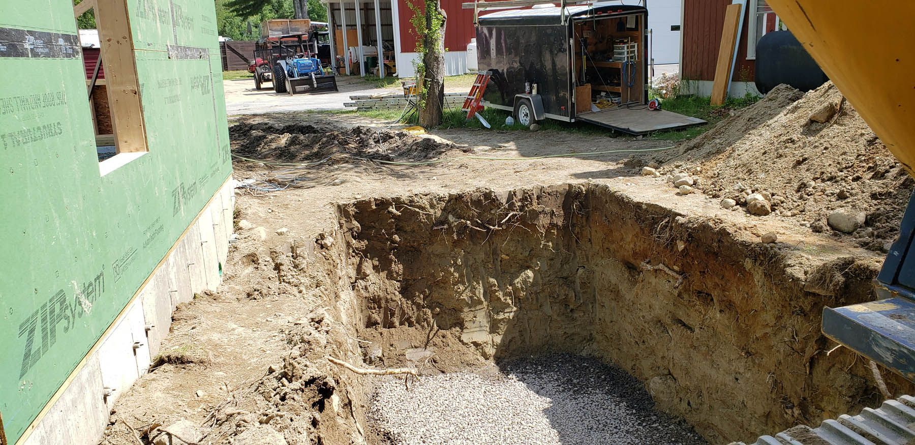 Excavation pit next to a green construction wall, with a concrete base and other construction equipment in the background.