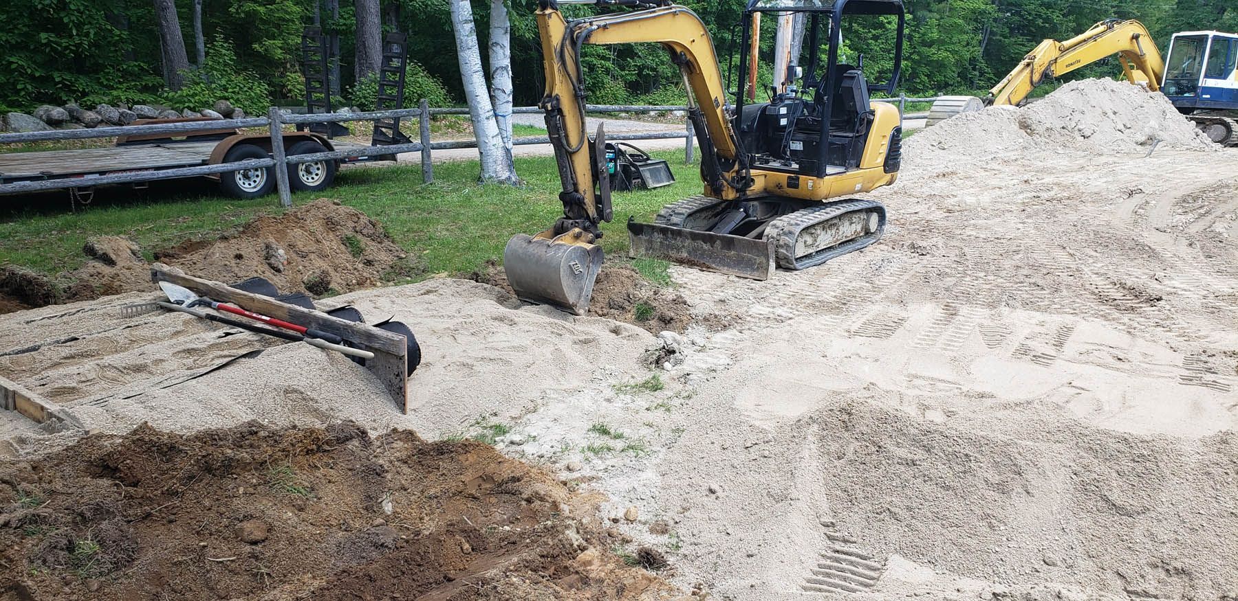 Yellow excavator digging in a gravel pile, trees in the background. Another excavator behind.