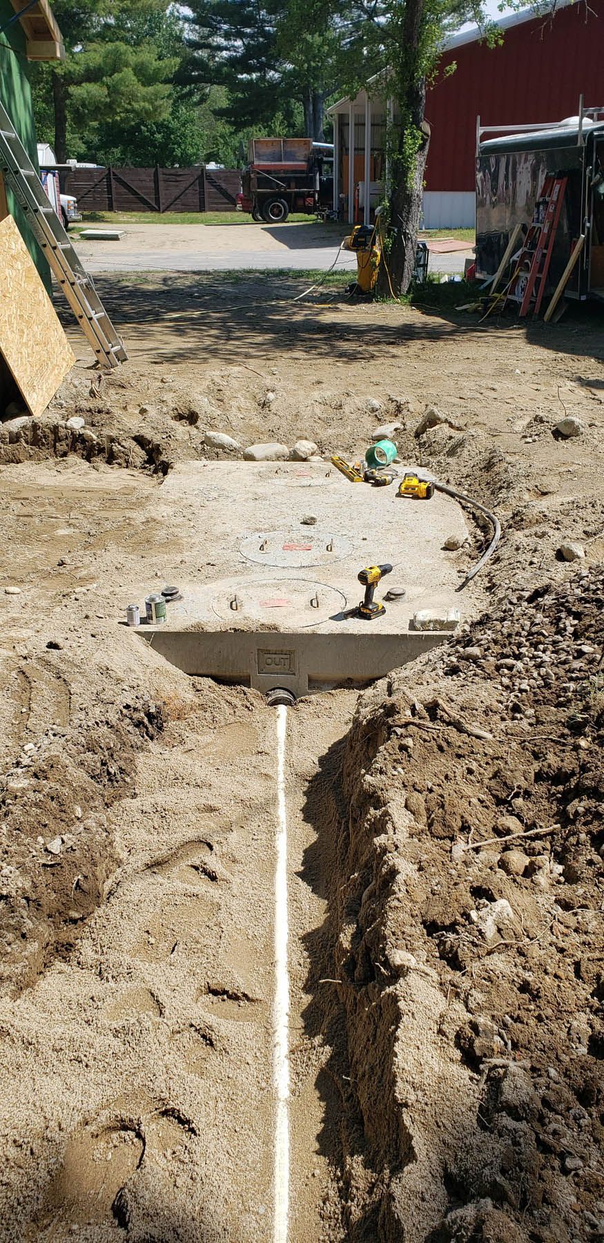 A buried concrete septic tank with a white pipe in a trench. Brown soil and a red barn.