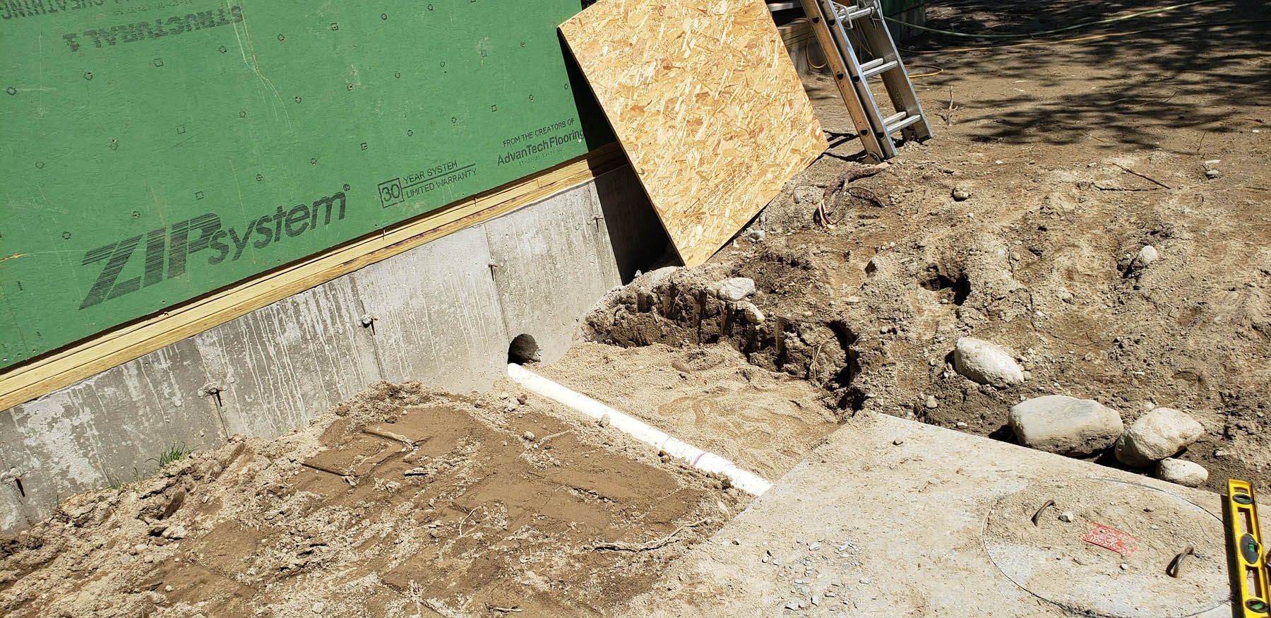 Construction site: foundation with green Zip System sheathing, exposed soil, PVC pipe, and wood panel.