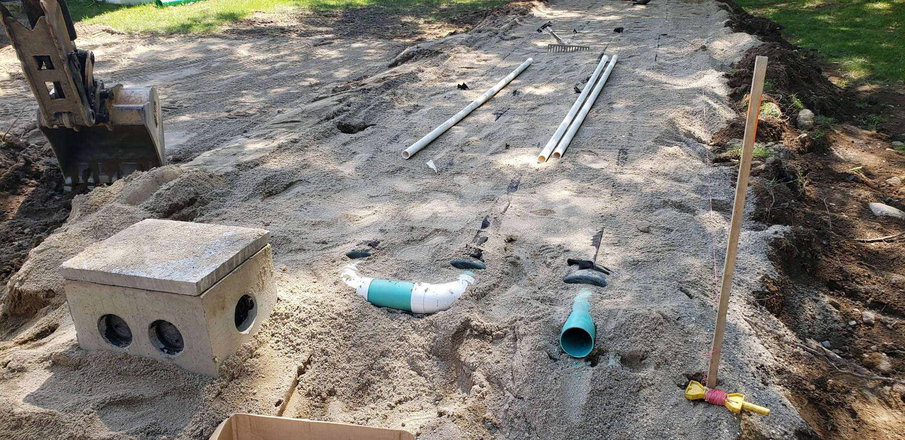 Construction site with a concrete box, pipes, and dirt. An excavator is in the background.
