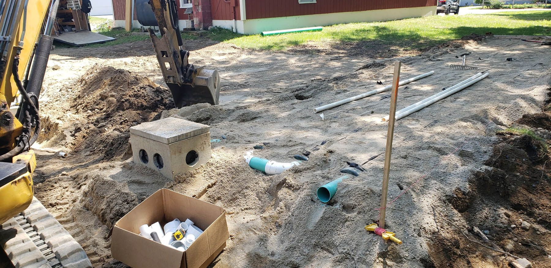 An excavator digging on a construction site. Pipes, a box of fittings, and a concrete structure are nearby.