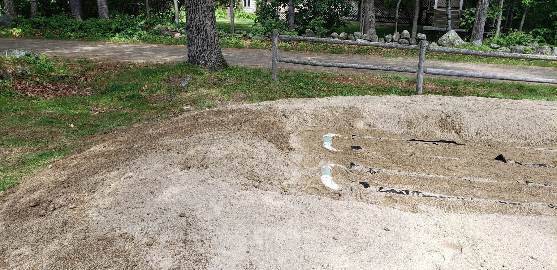 A sand pit with white drainage pipes, in a yard with a tree and grassy areas.